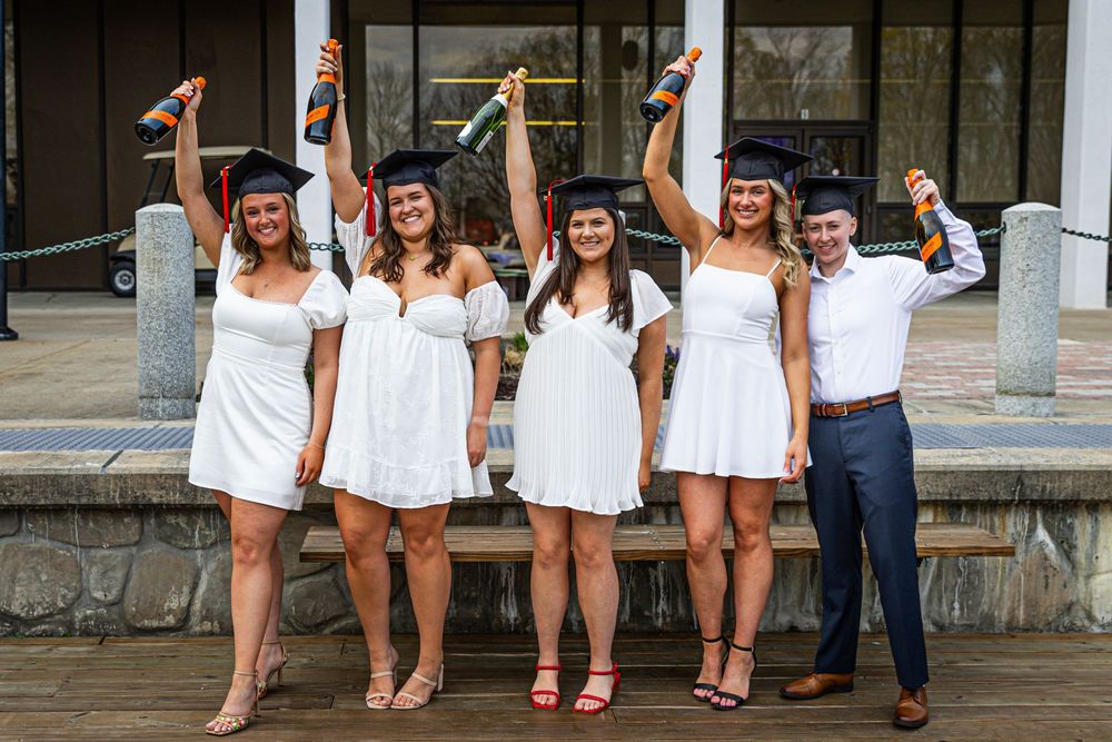 Five graduates in white dresses and caps celebrating outdoors holding diplomas in the air.