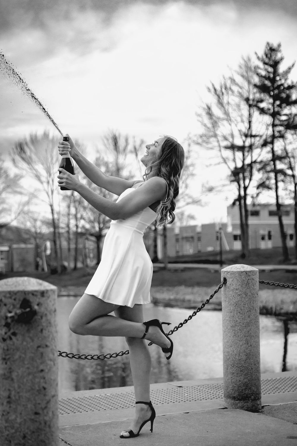 Woman in white dress popping champagne bottle outdoors near waterfront in black and white photo.