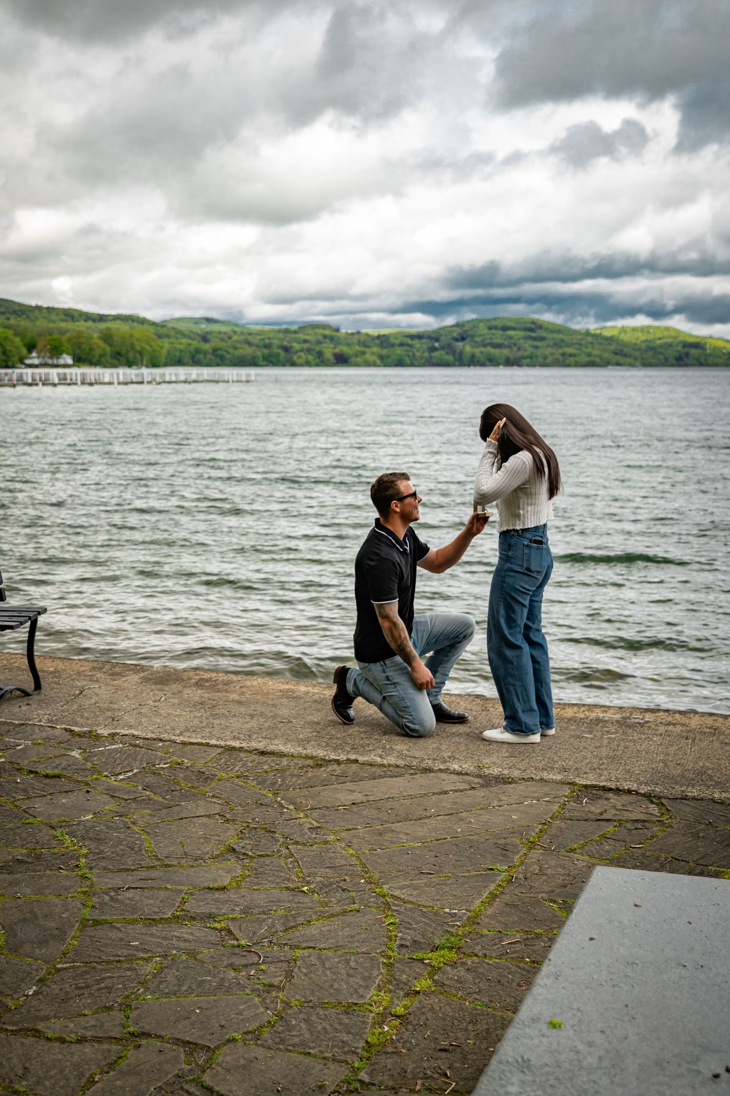 Man proposing on one knee to a woman by a lakeside on a cloudy day outdoors.