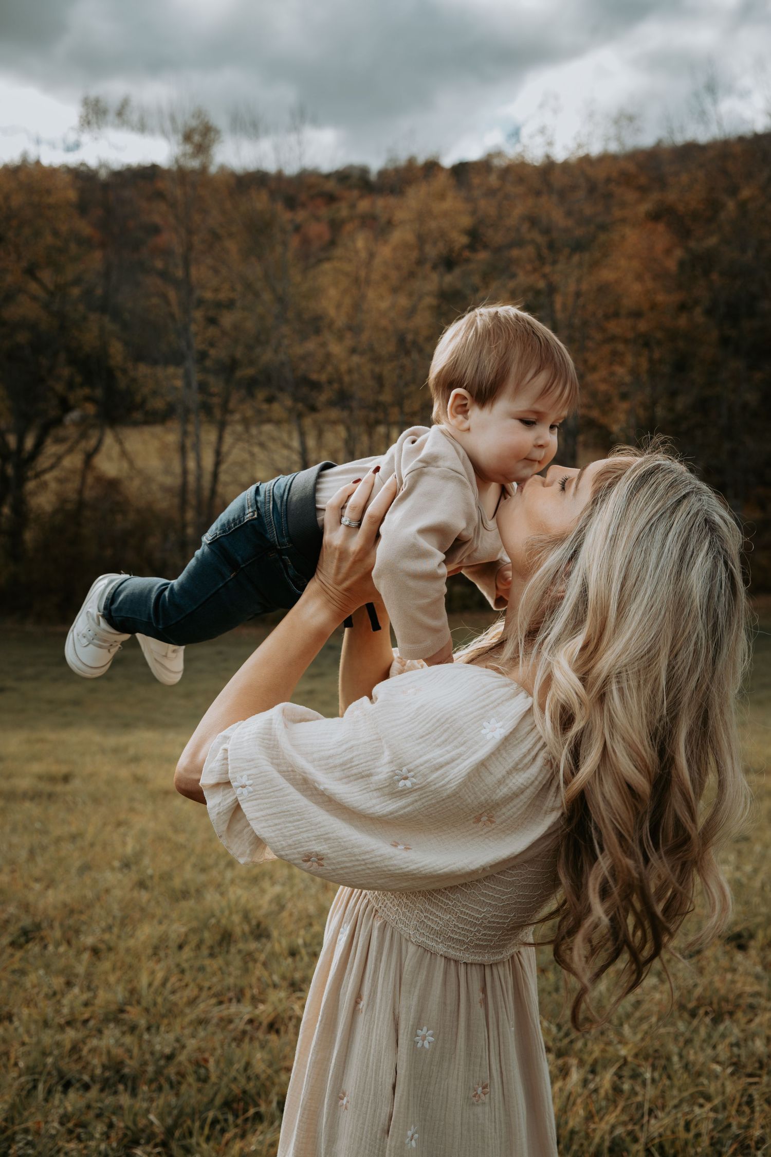 Mother lifting laughing toddler in the air against colorful autumn hillside backdrop.