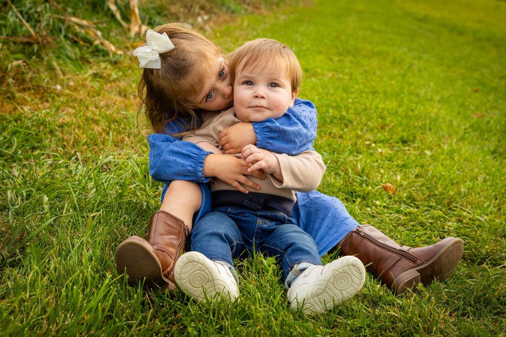 Two young siblings sitting on green grass, older girl hugging baby brother from behind.