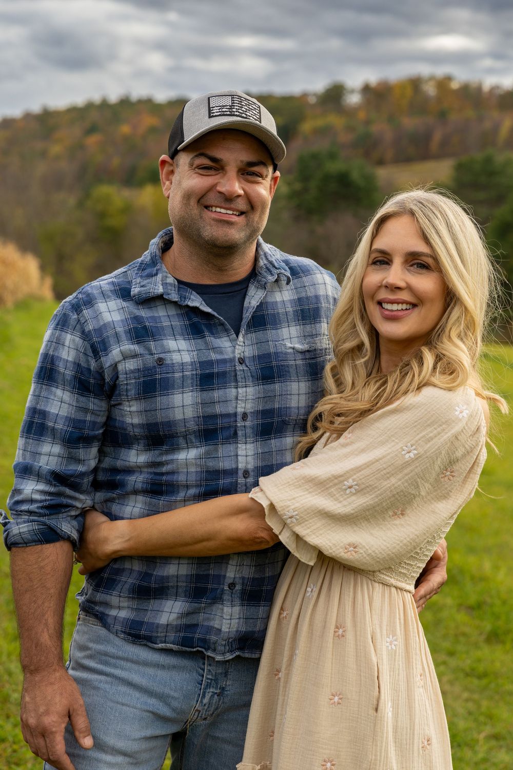 Smiling couple embracing outdoors in autumn landscape, man in plaid shirt and woman in cream dress.