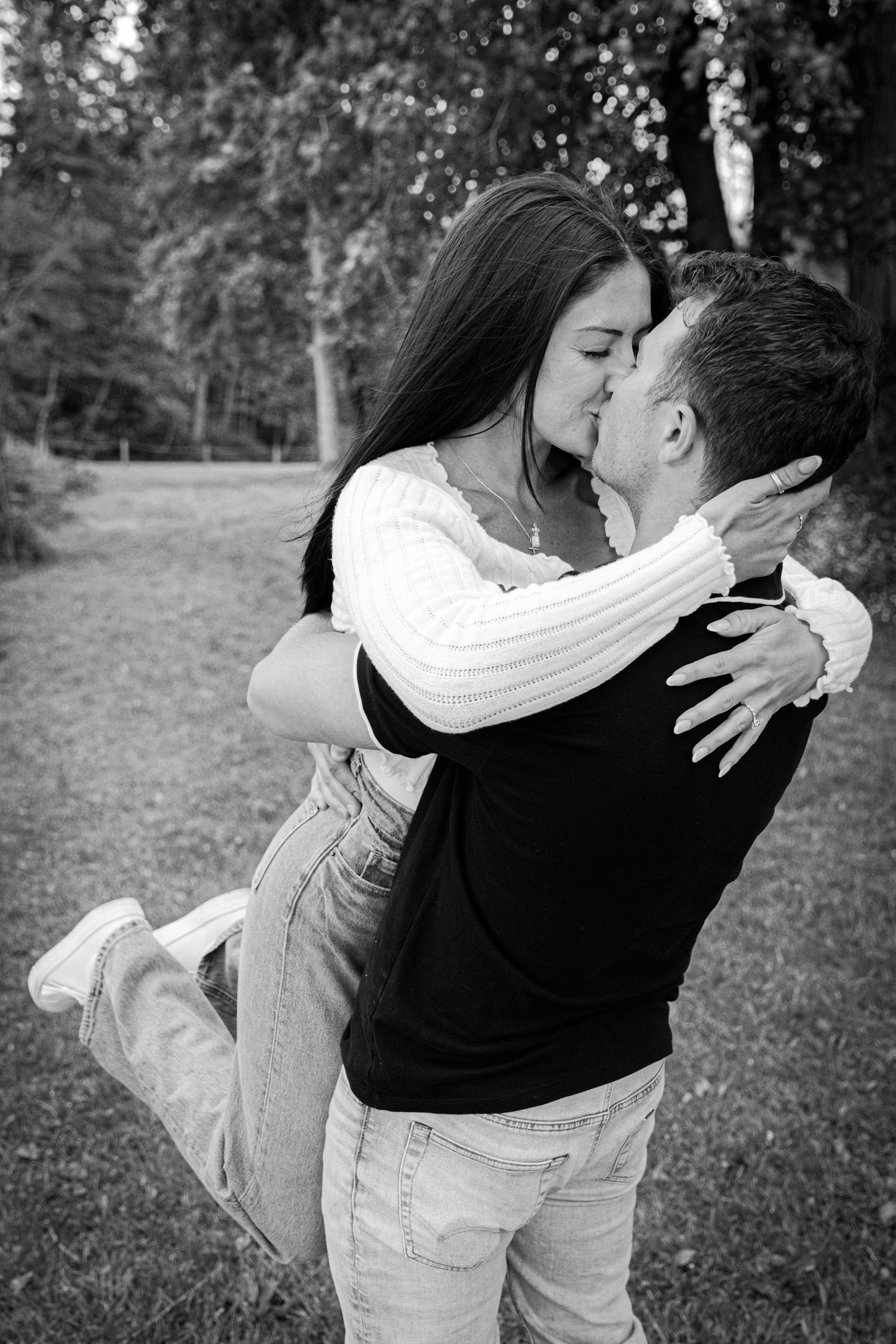 Black and white photo of a couple kissing and embracing in a park surrounded by trees.