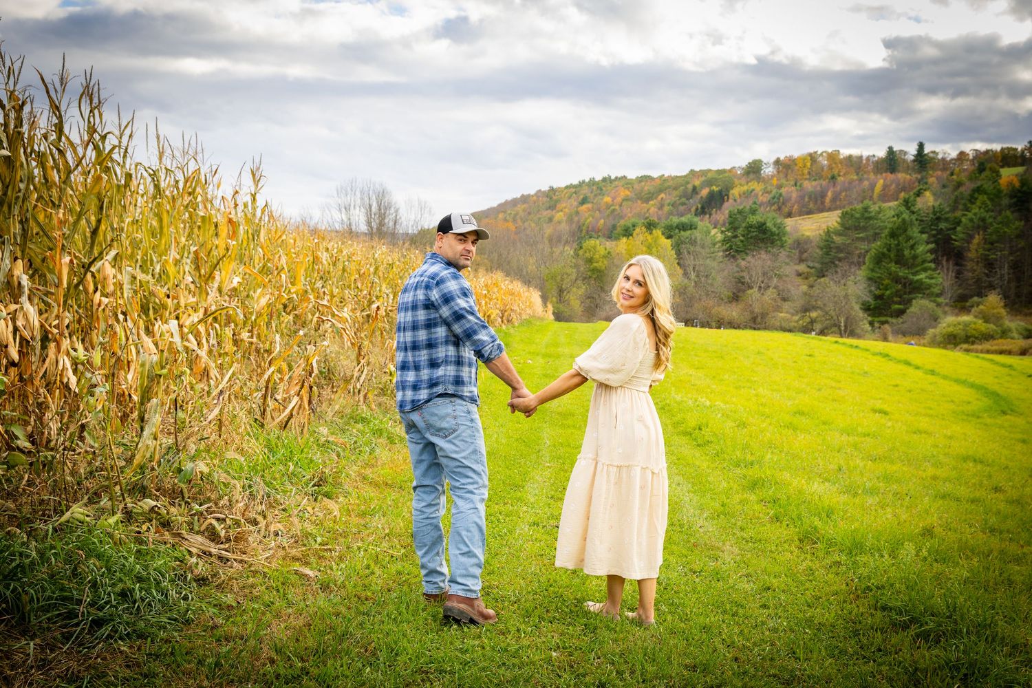 Couple holding hands near a golden cornfield and rolling hills during a fall outdoor photo session.