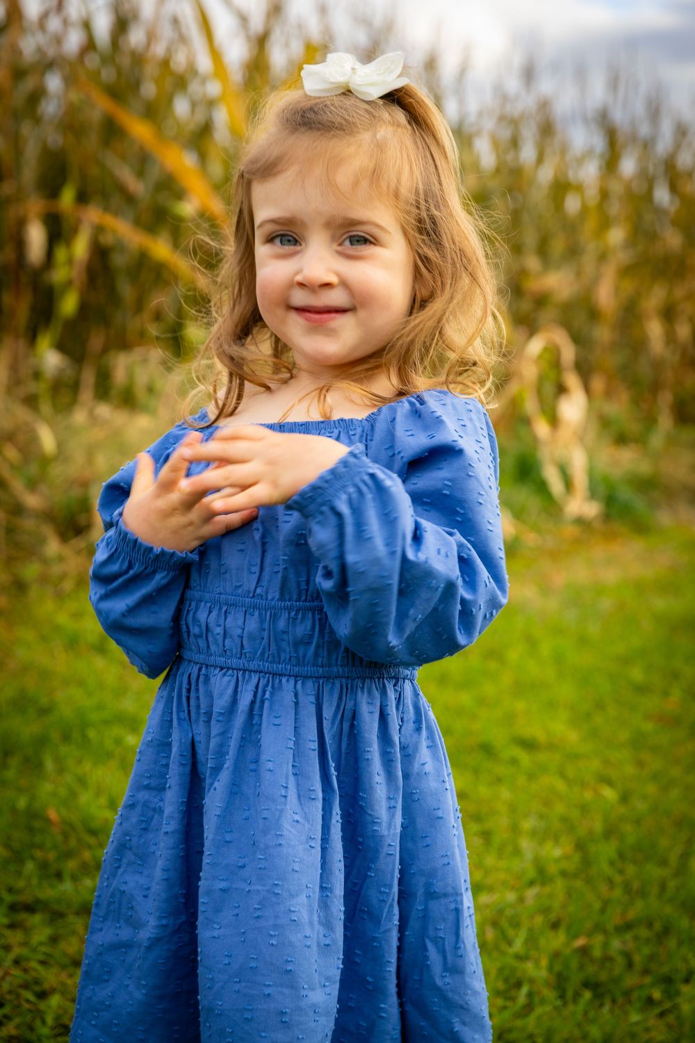 Adorable little girl in blue dress with white bow smiling in front of autumn cornfield.
