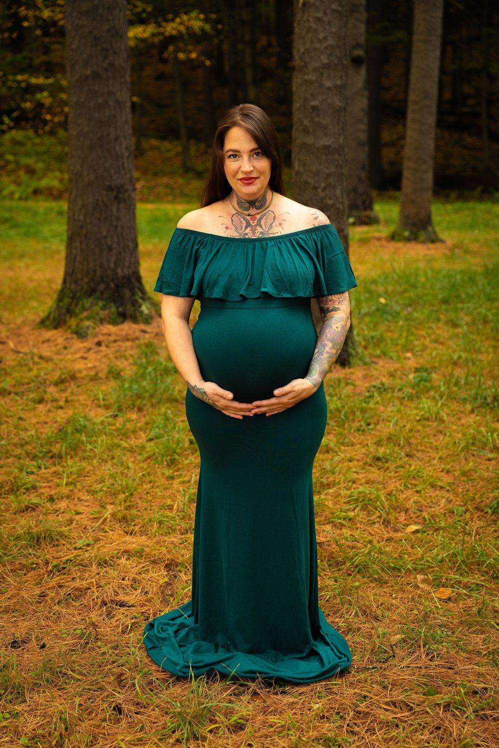 Pregnant woman in an elegant green off-shoulder gown cradling her baby bump in an autumn forest.