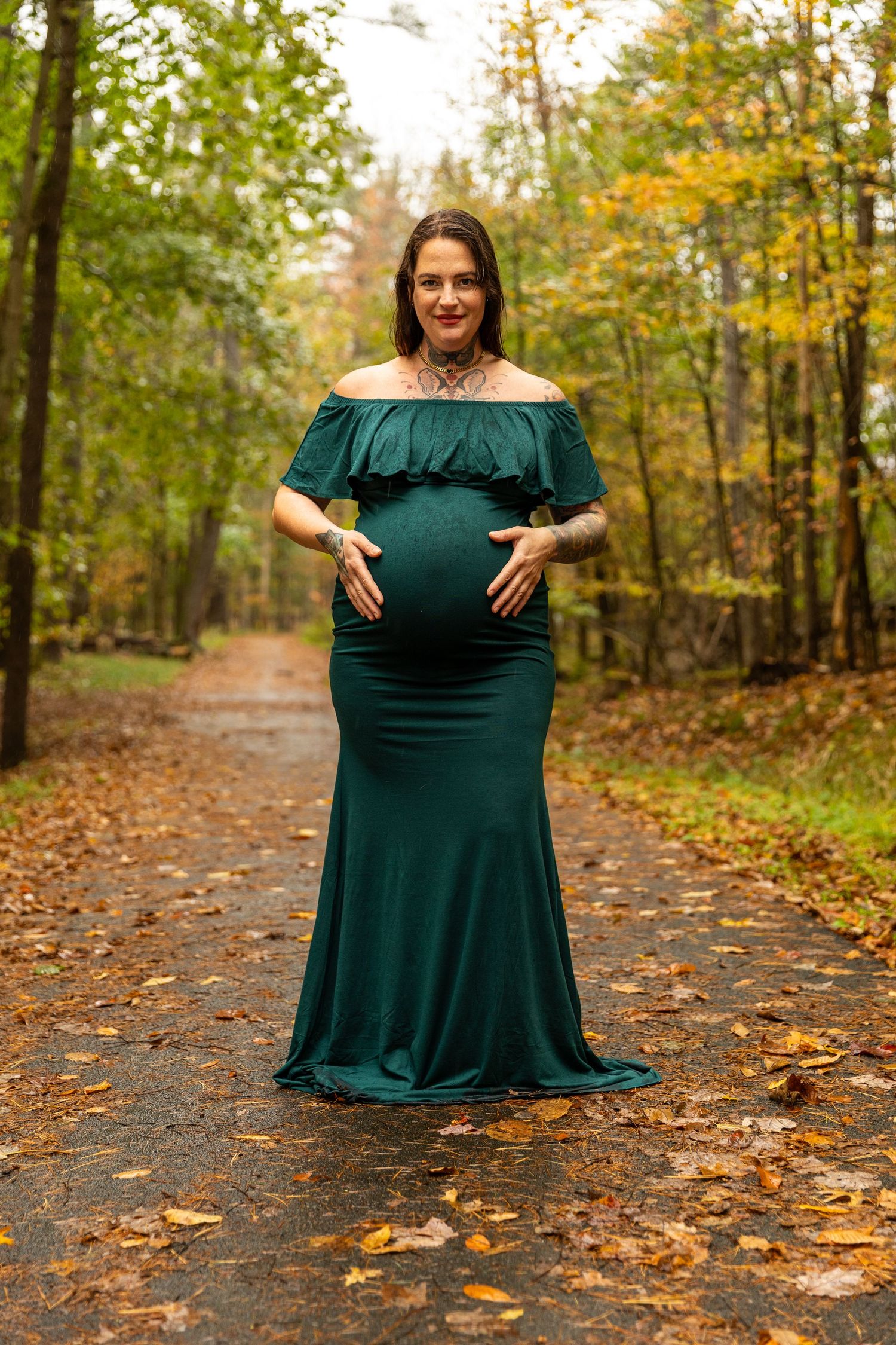 Pregnant woman in a teal off-shoulder gown standing on a leaf-covered path in an autumn forest.