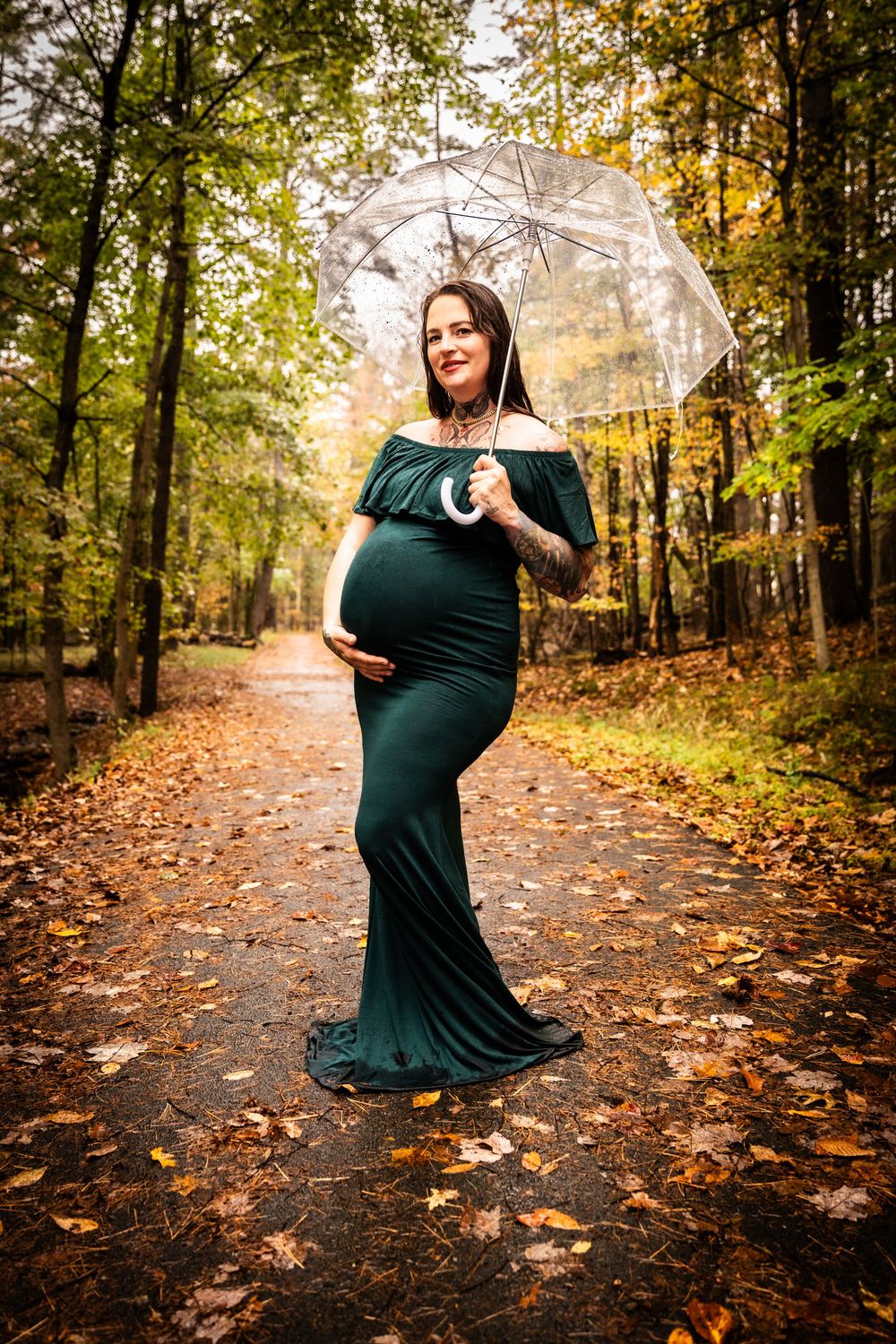 Pregnant woman in a teal gown holding a clear umbrella on a leaf-covered path in autumn forest.