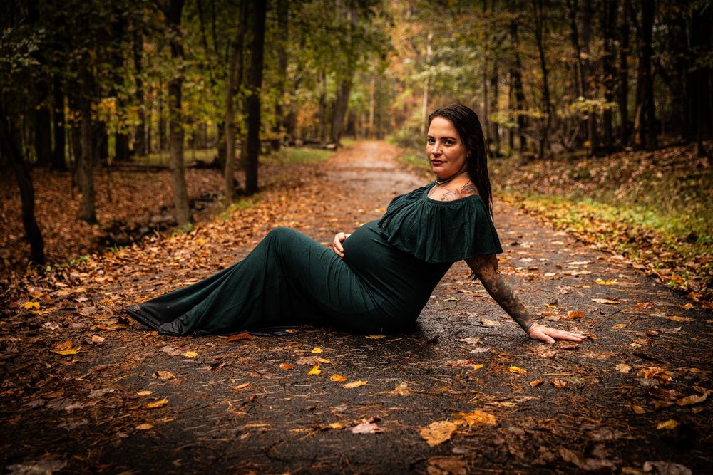 Pregnant woman in elegant green gown reclines on autumn forest path surrounded by fallen leaves.