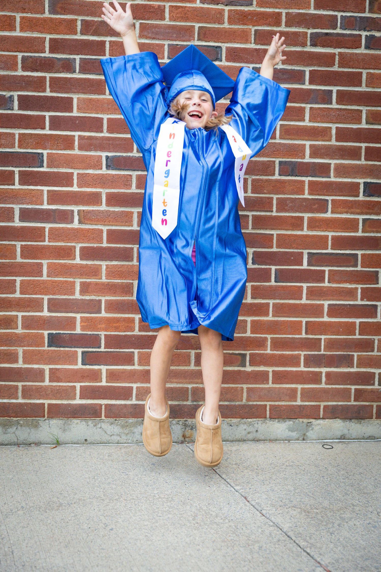 Excited young girl jumps for joy in blue graduation cap and gown against brick wall.