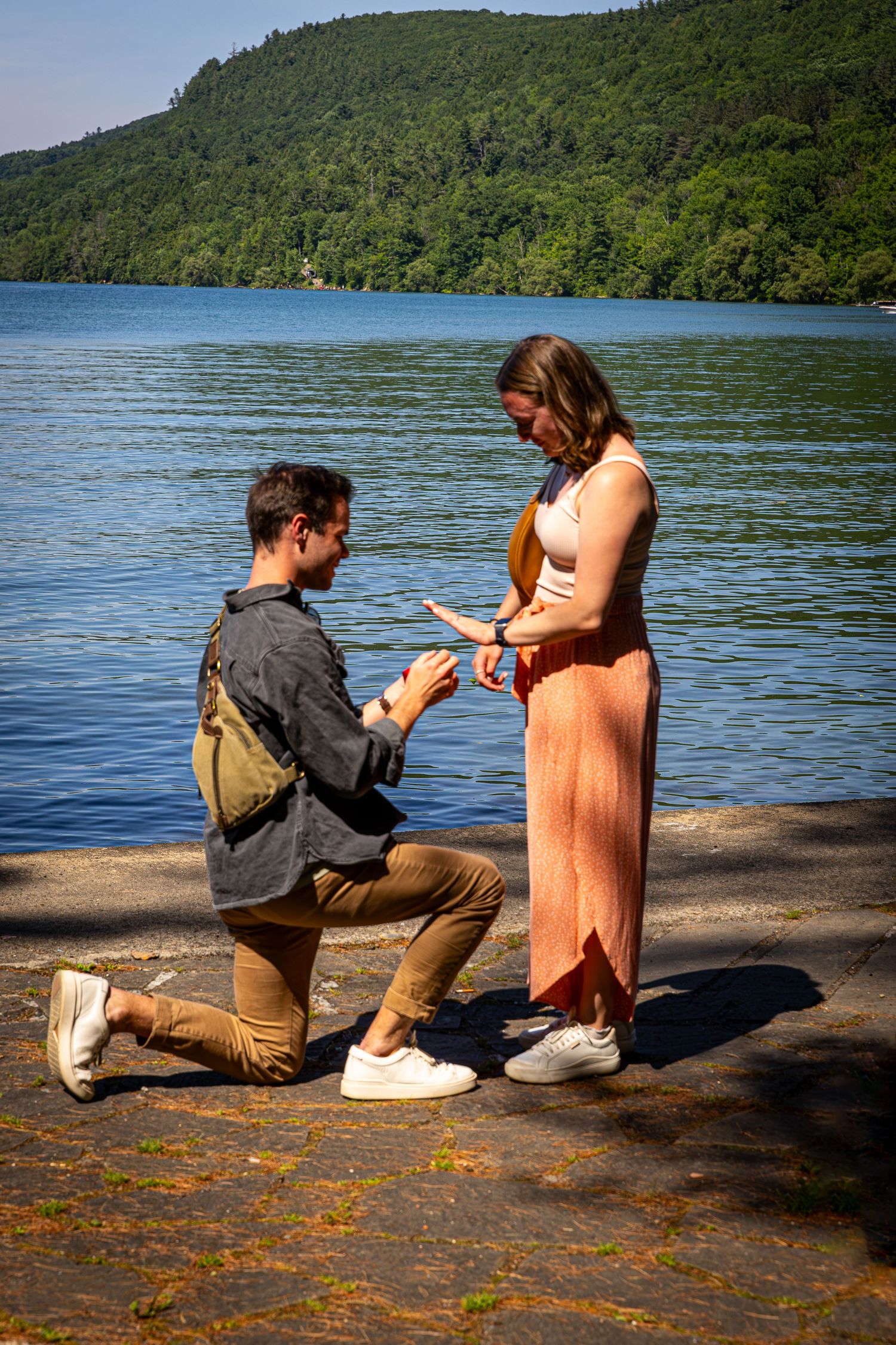 Man proposing on one knee to a woman in a pink dress beside a scenic blue lake on a sunny day.