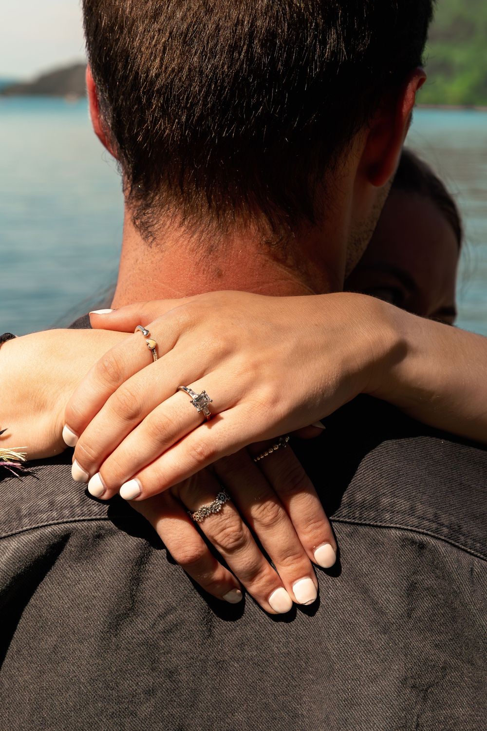 Close-up of a woman's hands with elegant rings embracing a man's shoulders near a serene waterfront.