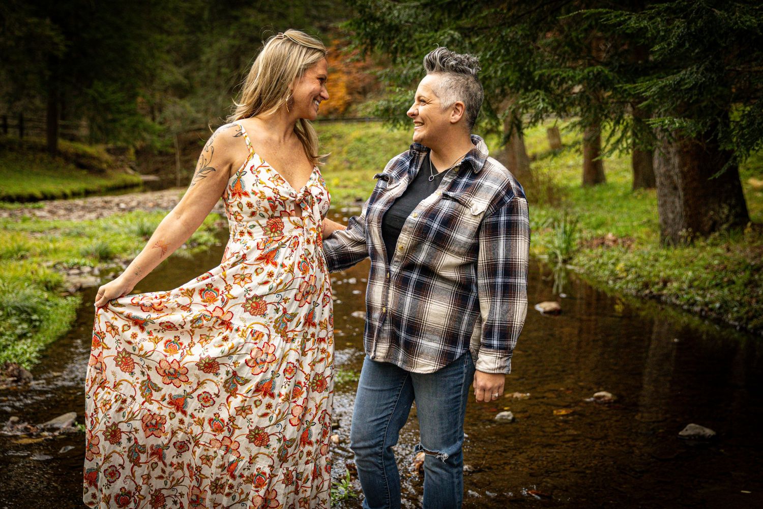 Smiling couple walking together beside a stream surrounded by lush green trees in autumn.