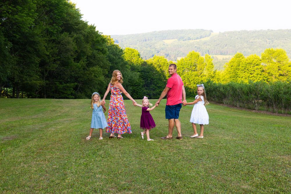 Family of five holding hands in a green meadow surrounded by vibrant trees on a sunny day.