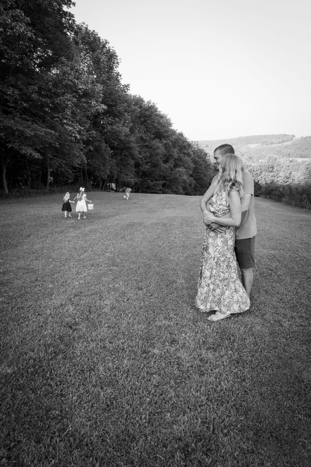 Black and white photo of couple embracing on grassy field while children play in background.