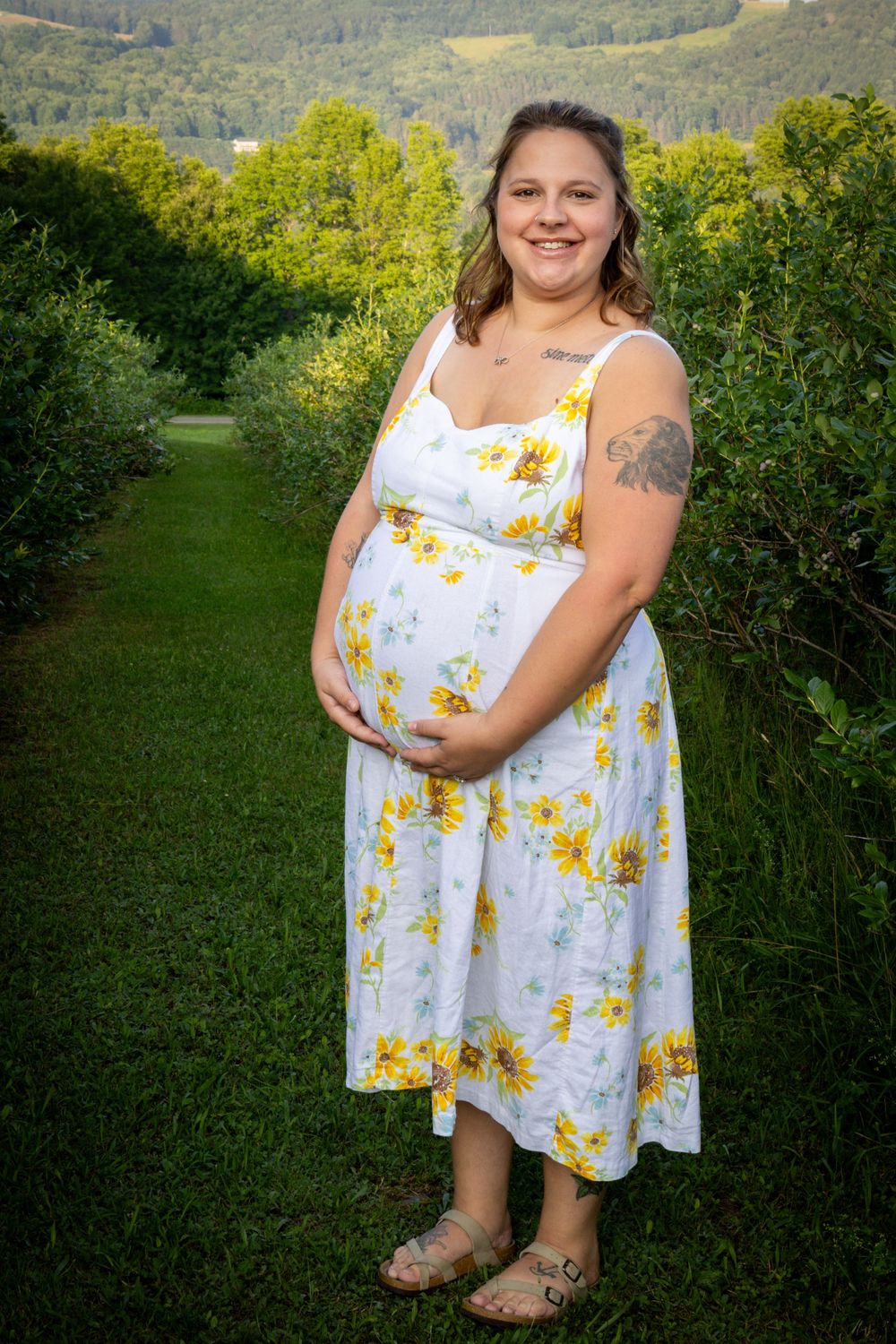 Smiling pregnant woman in white sunflower dress cradling baby bump in lush green garden.