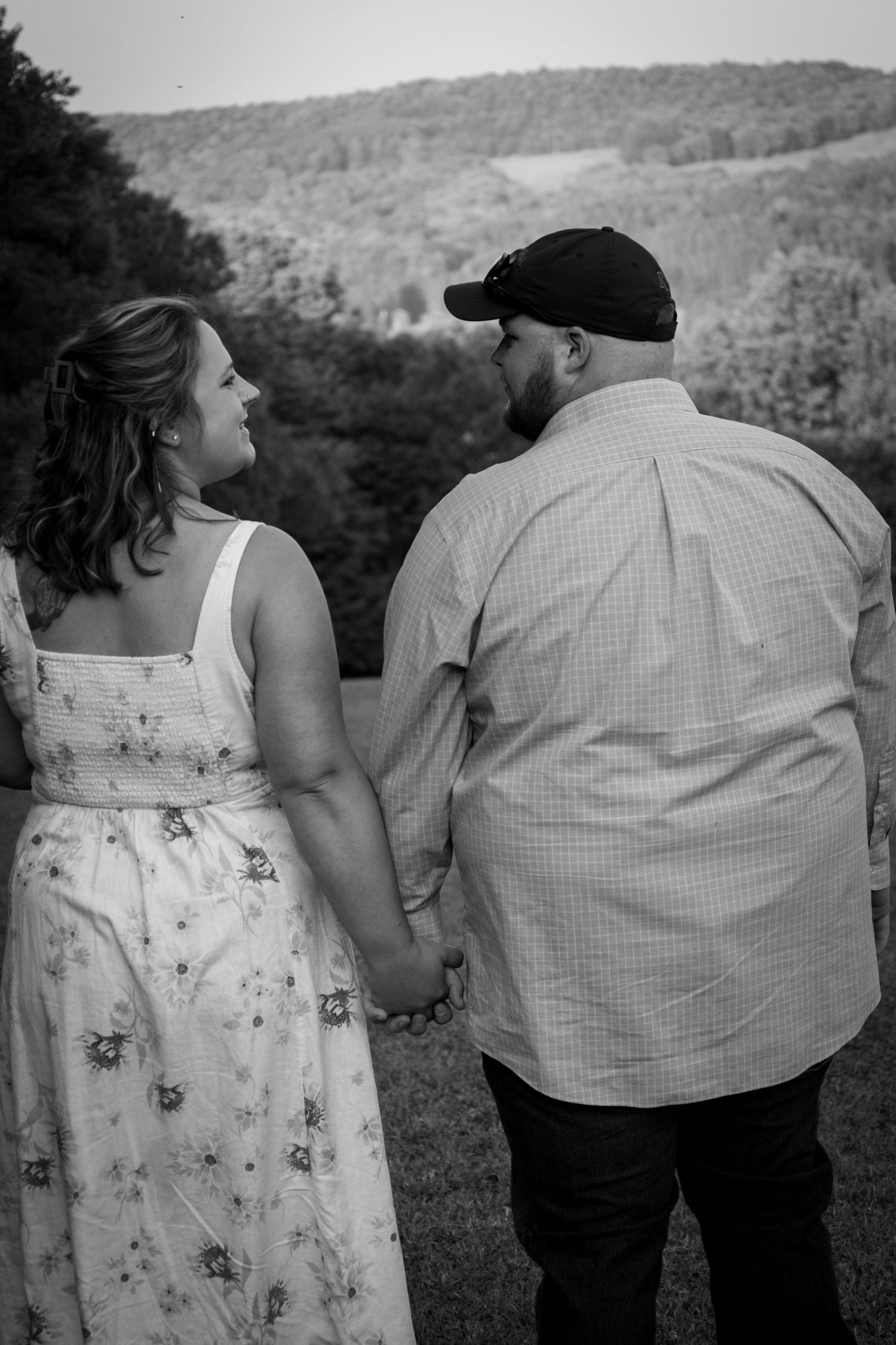 Black and white photo of pregnant couple holding hands while gazing at scenic hillside view.