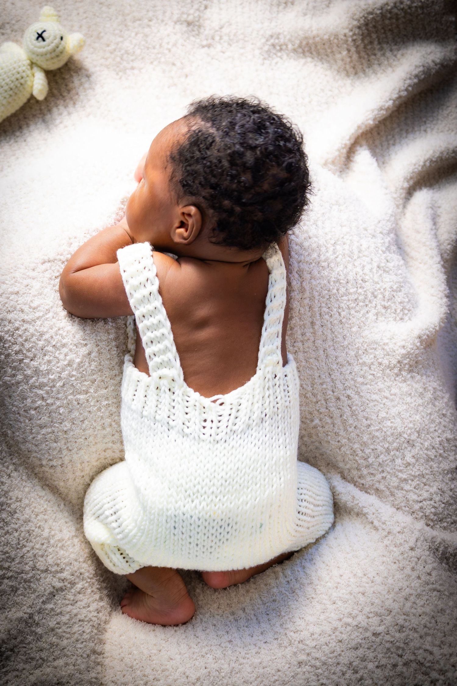 Sleeping newborn baby in white knit romper lying on soft blanket with stuffed toy nearby.