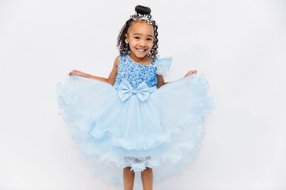 Adorable young girl twirls in a blue sequined tulle dress against a white background.
