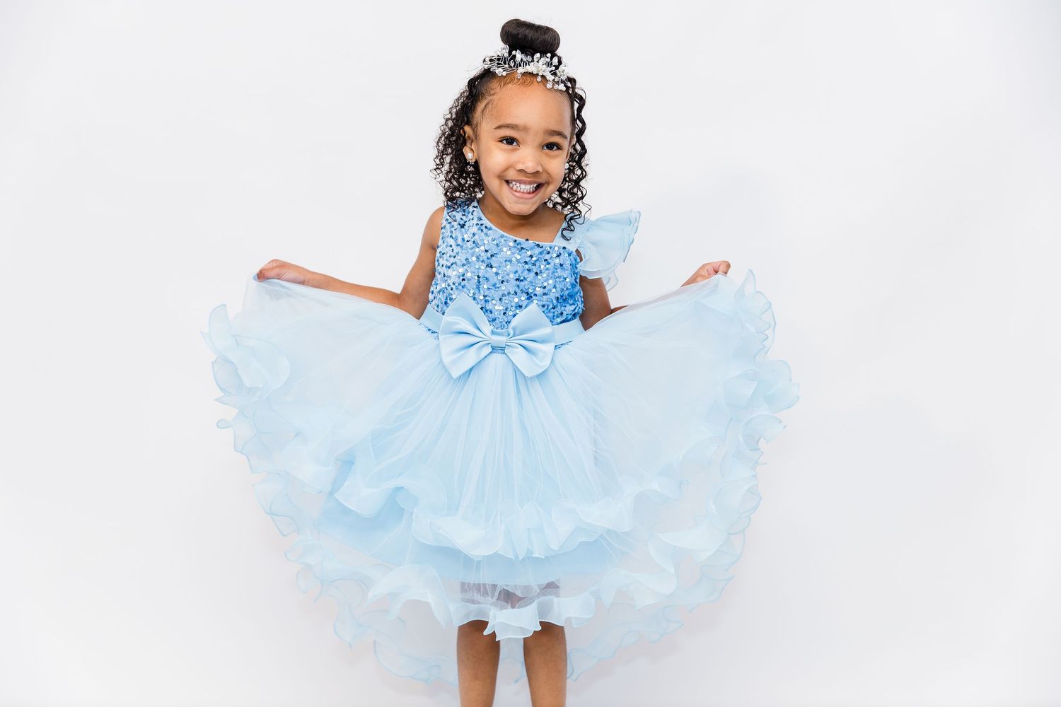Adorable young girl twirls in a blue sequined tulle dress against a white background.