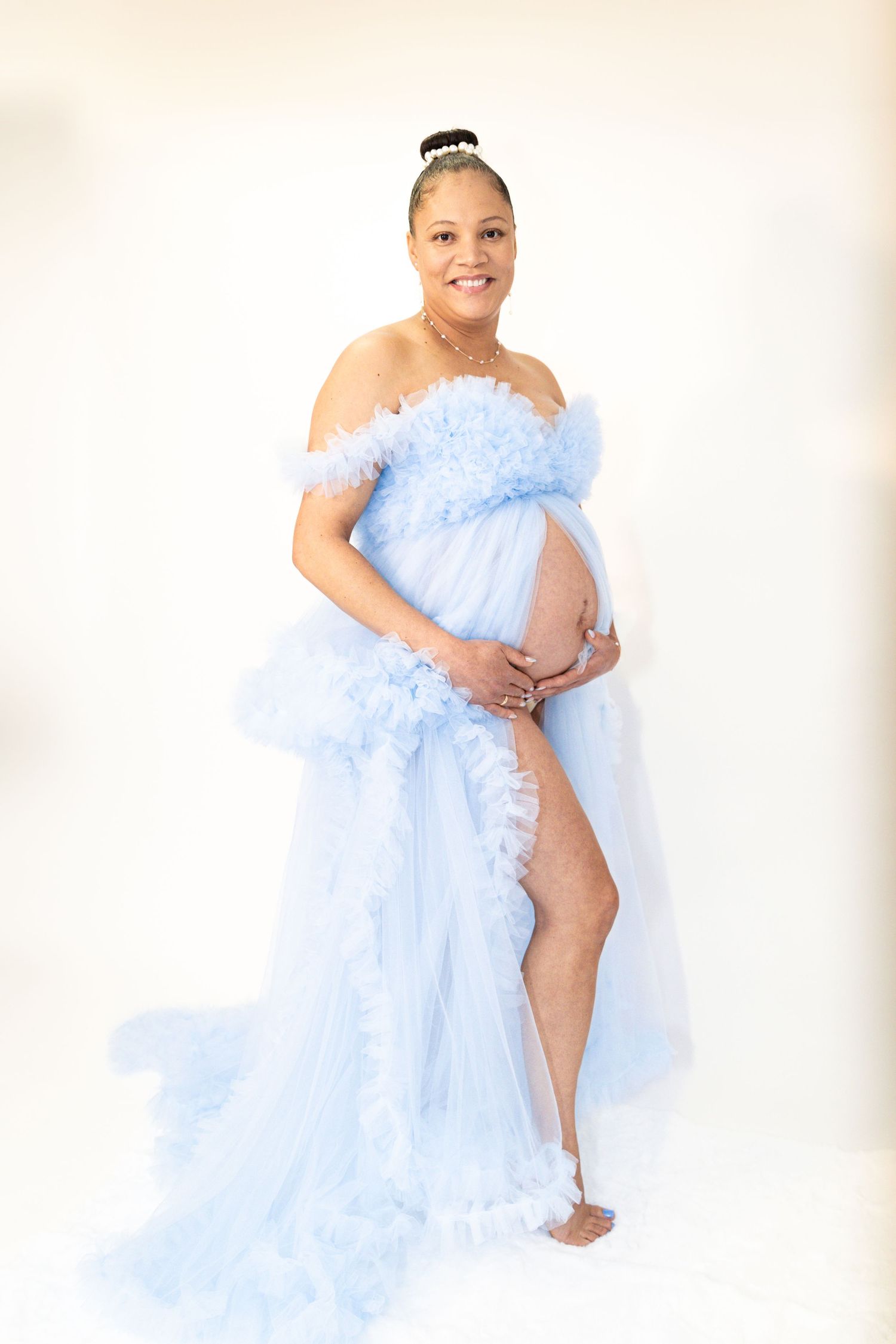 Radiant pregnant woman poses in a flowing light blue tulle maternity gown on white background.