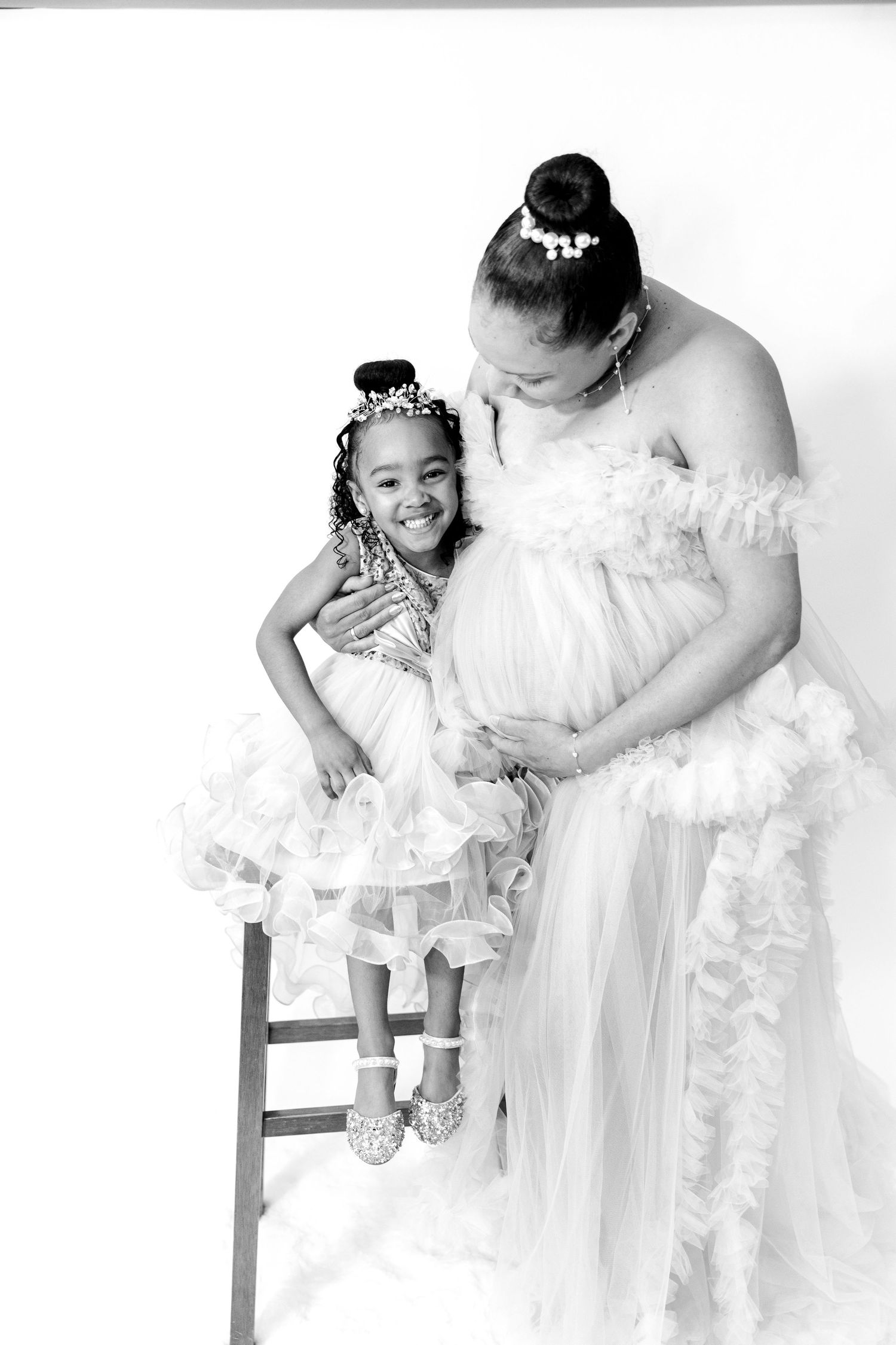 Black and white portrait of a pregnant woman in white gown tenderly embracing her young daughter.