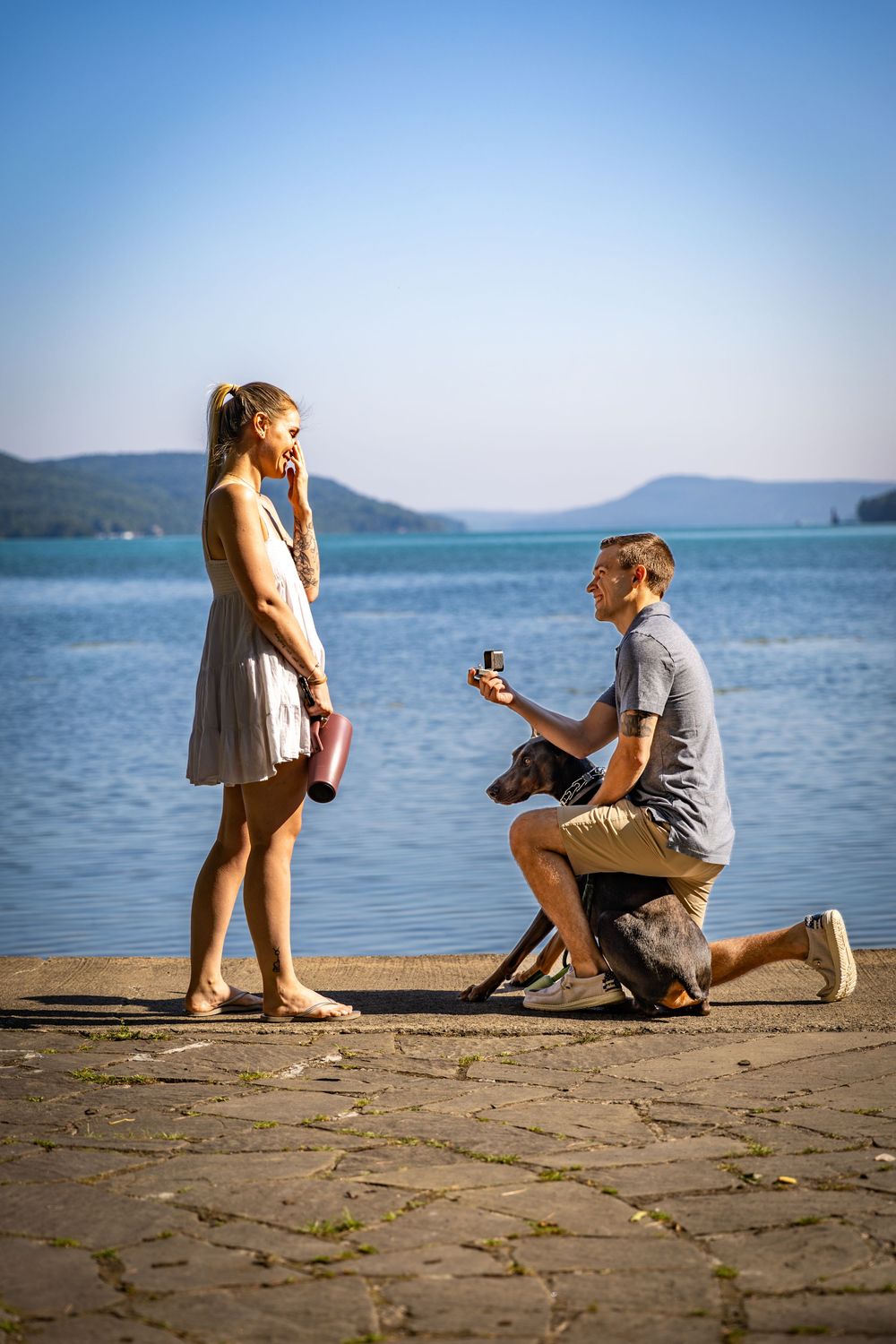 Man proposes on one knee to his girlfriend by a serene lake with their dog nearby.