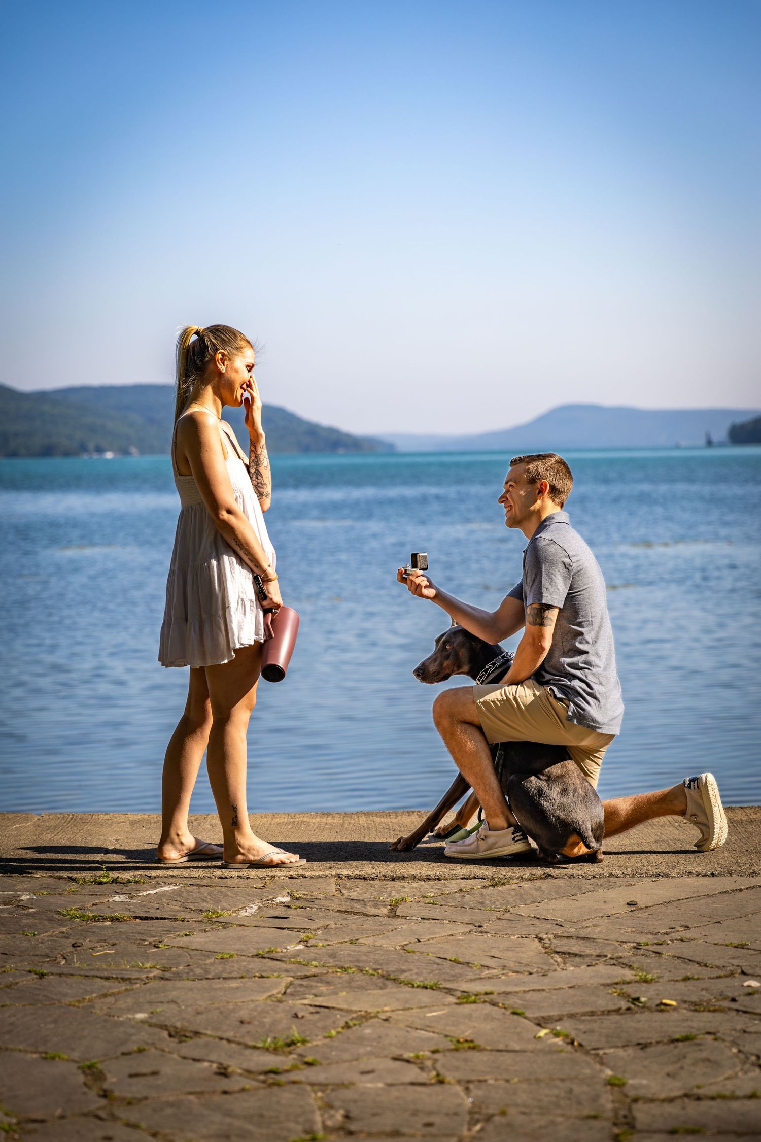 Man proposes on one knee to his girlfriend by a serene lake with their dog nearby.