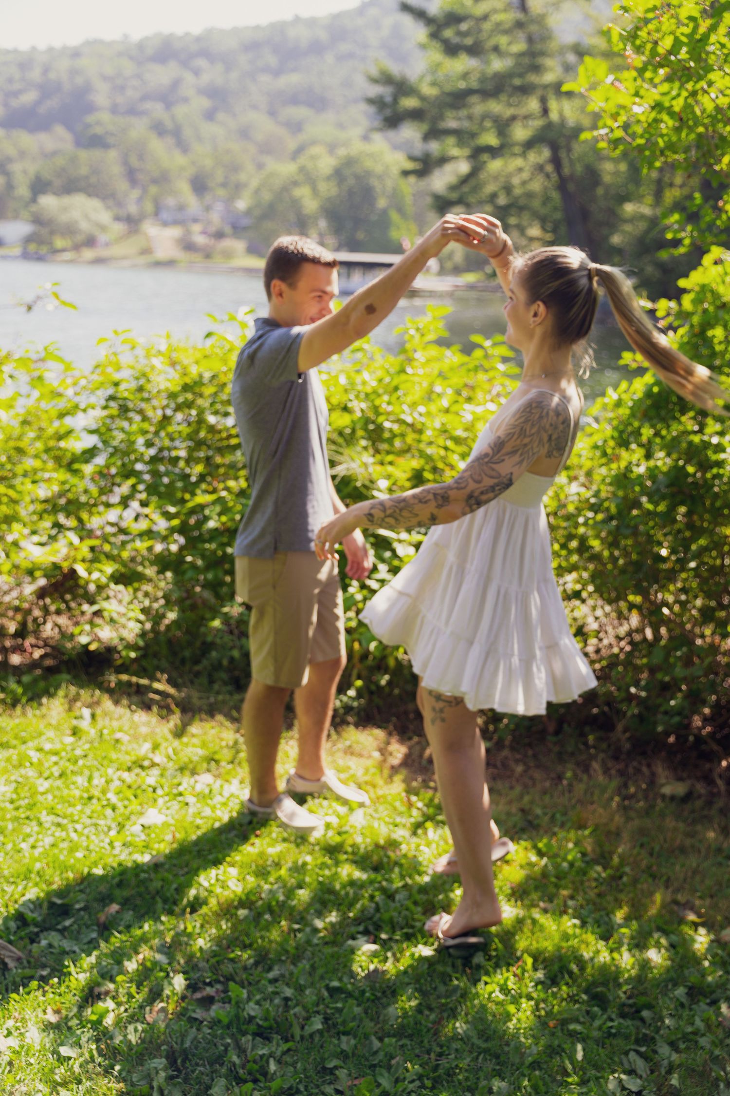 Happy couple dances together on a sunny green lawn with a scenic lake in the background.
