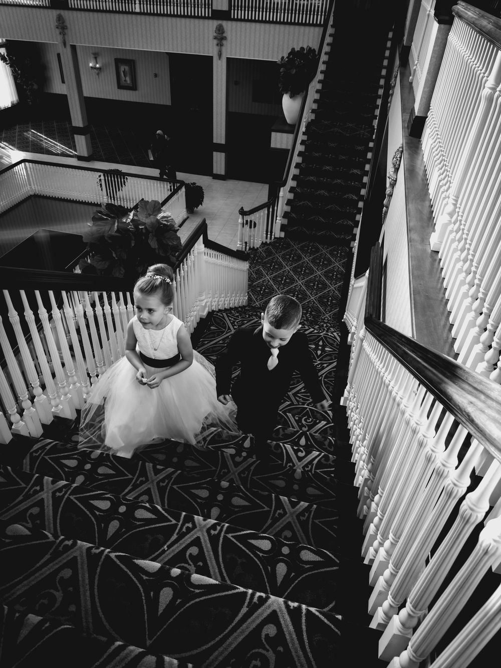 Black and white aerial view of flower girl and ring bearer walking down grand staircase at wedding venue.