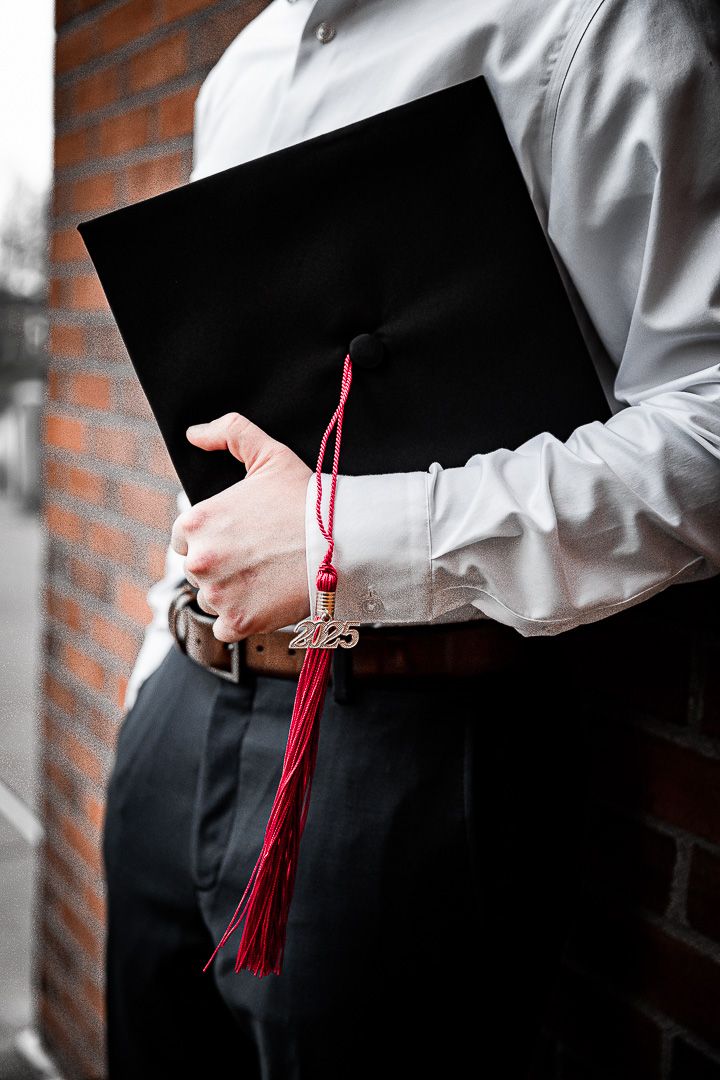 Graduate holding a black mortarboard cap with a red 2025 tassel against a brick wall background.
