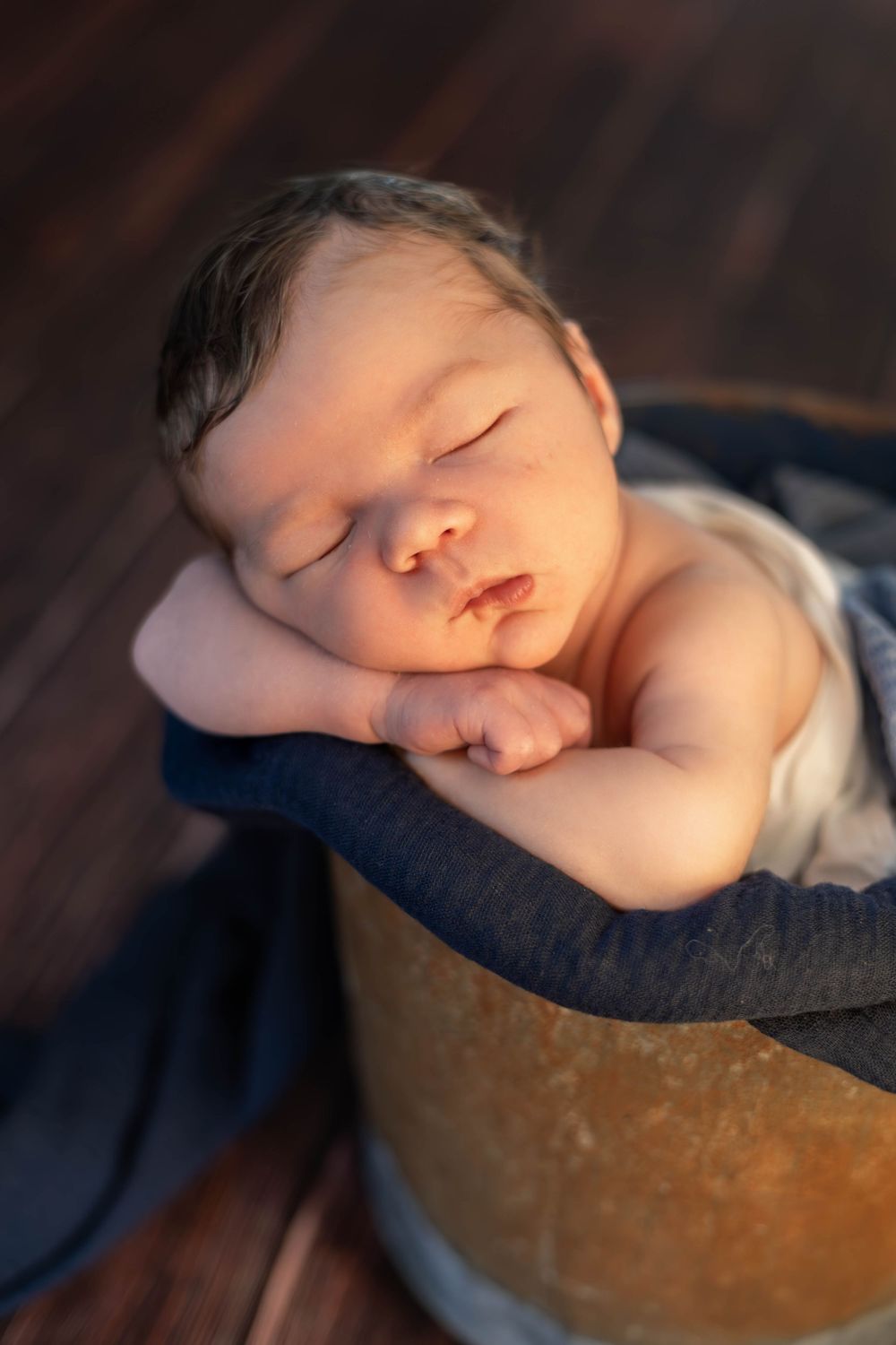A sleeping newborn baby rests peacefully in a dark blue blanket against a shadowy background.