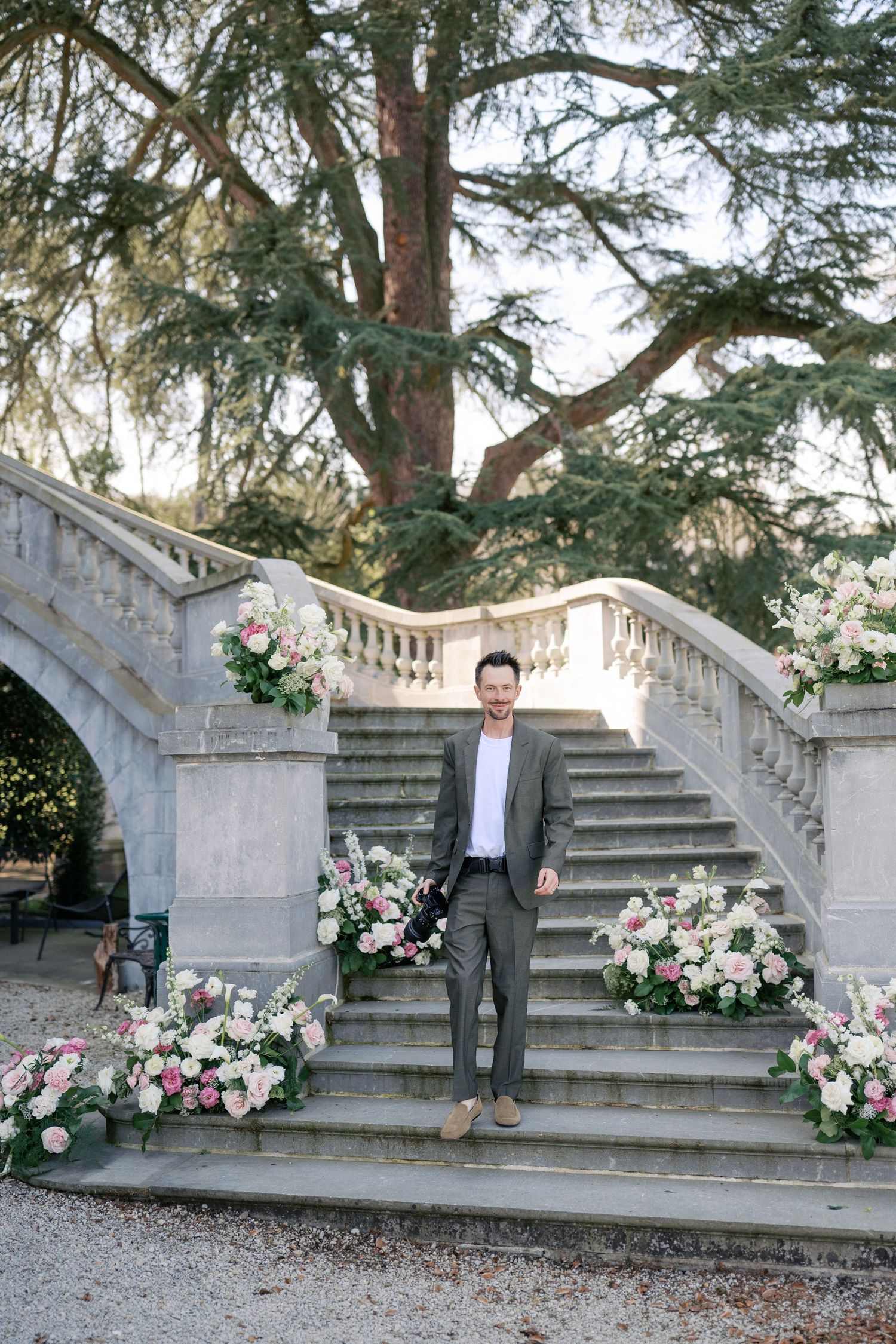 A person in a beige suit poses on stone steps outside a Mediterranean-style building with cypress trees.