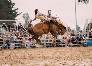 A saddle bronc rider is airborne above a bucking horse in a dirt arena as rodeo fans watch from the grandstand.