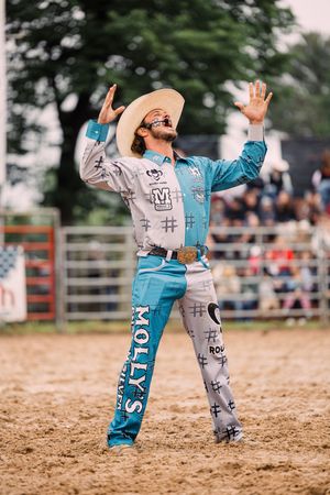 A rodeo bullfighter in a colorful sponsor-patched outfit and cowboy hat raises both hands to the sky in the arena.