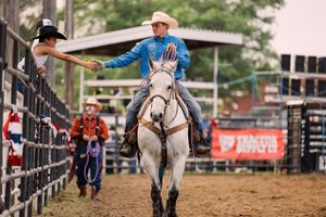 A cowboy on a gray horse reaches out to shake hands with a person at the fence while rodeo hands wait in the arena.