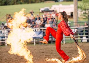 A performer in a red outfit stands in the rodeo arena beside a tall burst of flames as spectators watch from the stands.