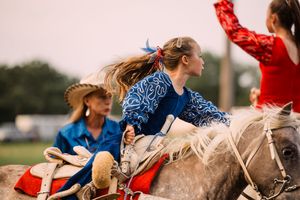 A young rodeo rider with a blue ribbon in her hair sits on a gray horse as another rider in red raises an arm nearby.