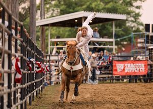 A trick rider in white stands tall on a galloping chestnut horse with one arm raised as the rodeo crowd watches from the stands.