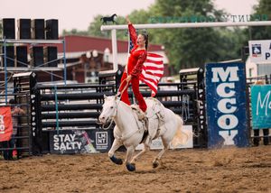 A rider in red waves an American flag while a white horse gallops across the rodeo arena past McCoy and Stay Salty banners.