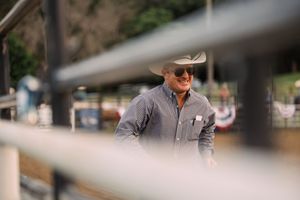 A smiling cowboy in a white hat, sunglasses and gray button-up shirt stands behind a blurred rodeo arena fence.