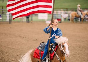 A young girl in a blue dress rides a palomino horse carrying a large American flag across a rodeo arena.