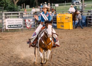 A smiling rodeo queen in a sash and cowboy hat rides a paint horse at a gallop and waves in a dirt arena.