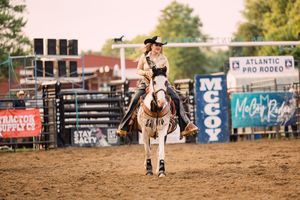 A rodeo queen in a cowboy hat rides a white horse across the dirt arena past McCoy and Atlantic Prodeo sponsor banners.