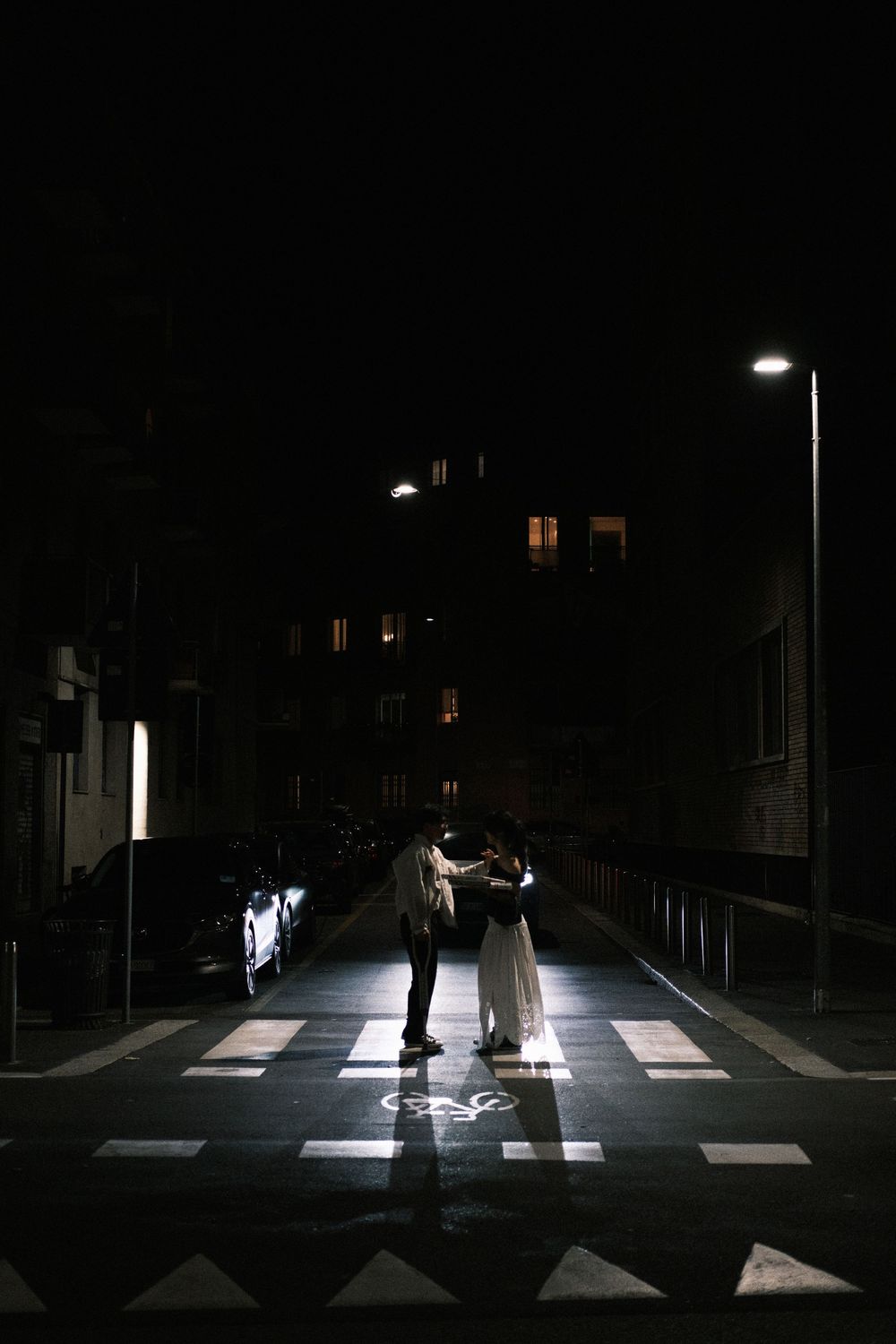 Pedestrians crossing an illuminated zebra crossing at night with street lights creating dramatic shadows.