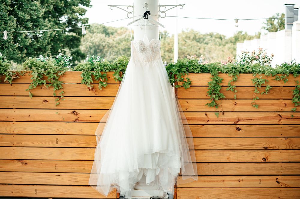 White wedding dress hangs elegantly against a rustic wooden fence backdrop with greenery.