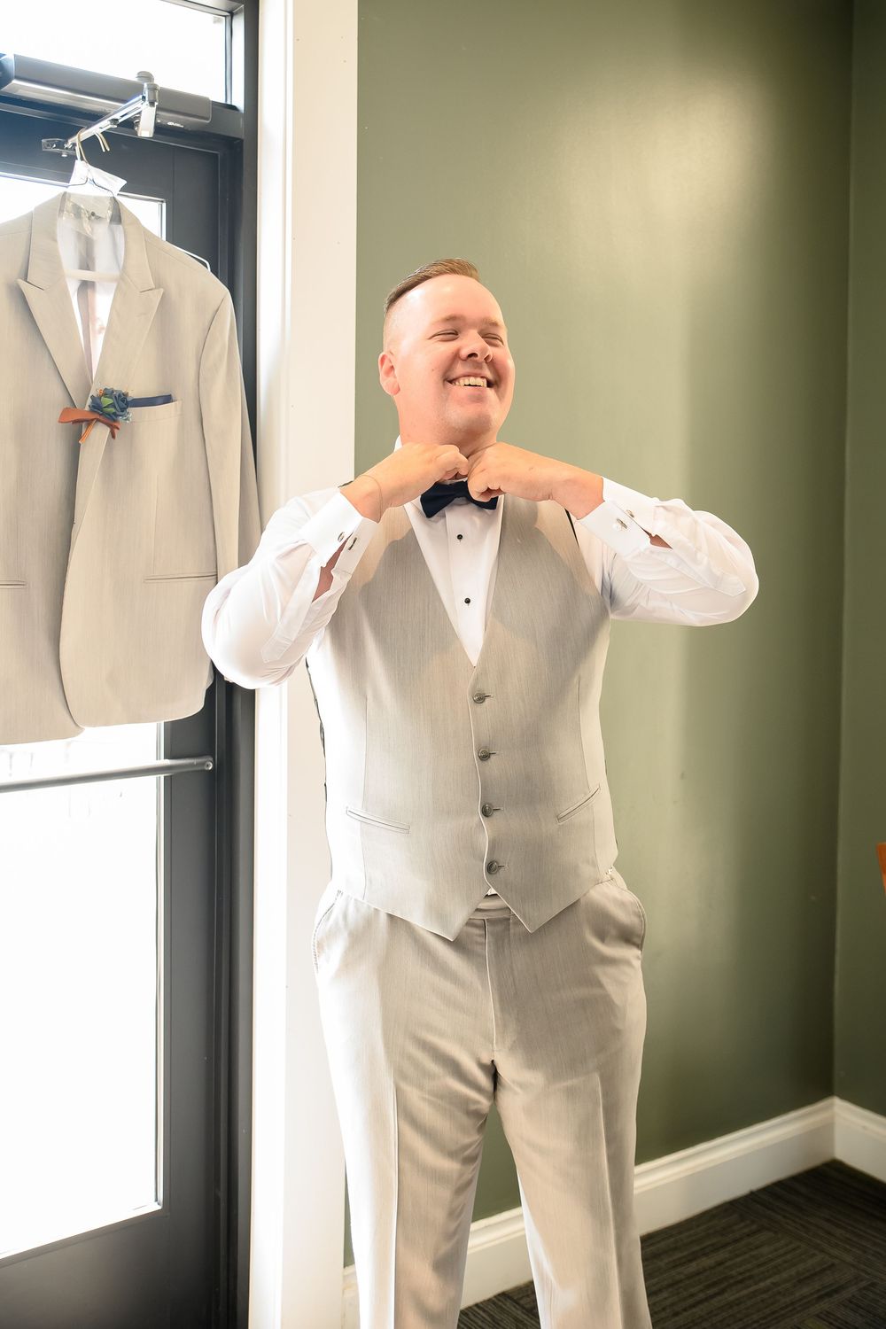 A person in a white formal suit and bowtie poses in a dressing room setting.