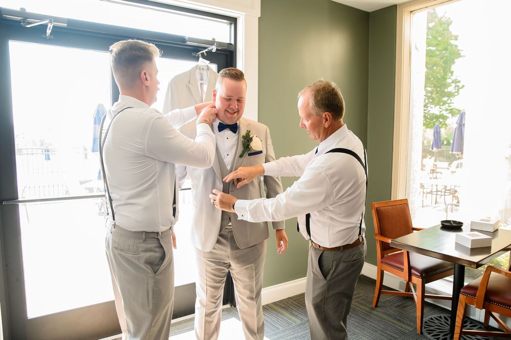 Wedding party members in light colored suits preparing in a room with large windows.