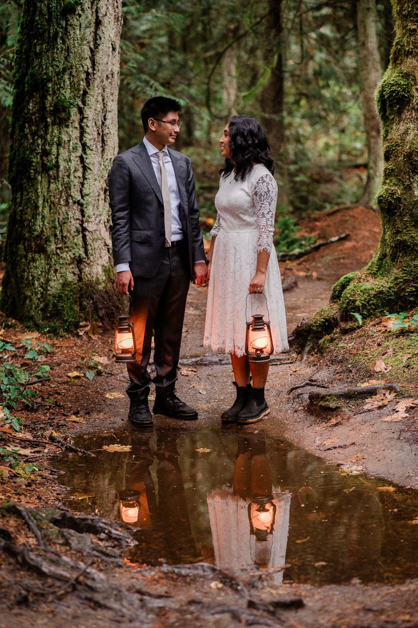 A couple holding lanterns stands on a forest path with their reflection in a puddle during Bainbridge Island Elopement