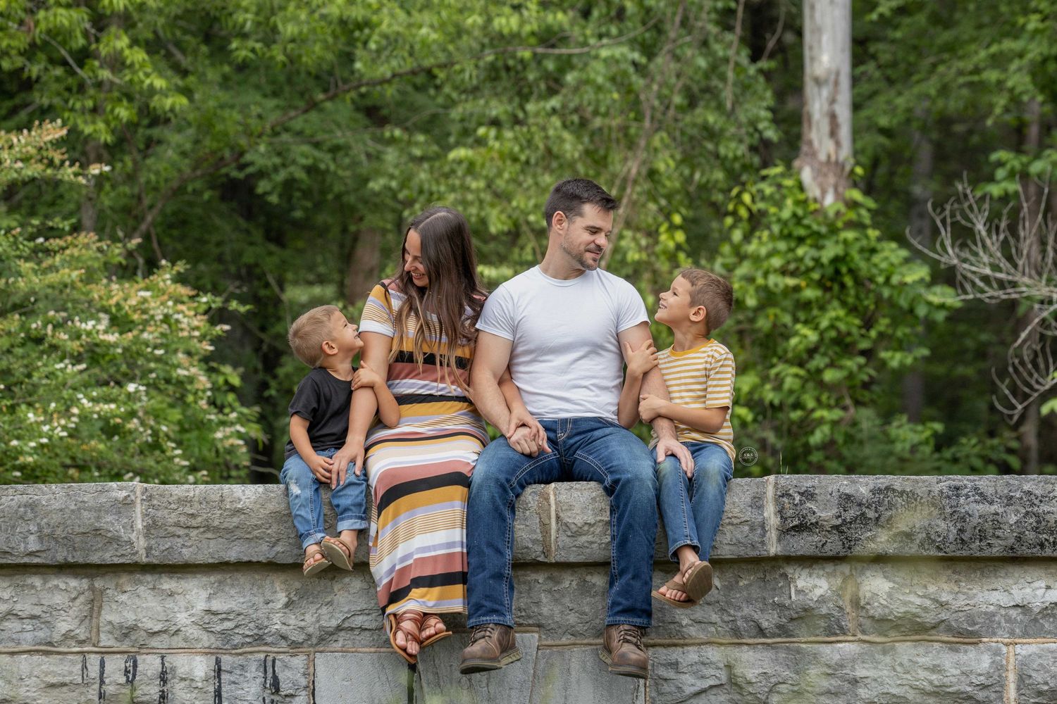 A couple sits together on a rustic stone wall surrounded by lush green foliage on a warm summer day.